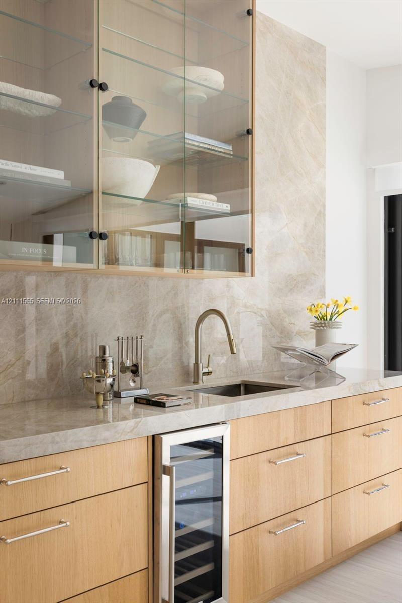 Wet bar featuring light-toned wood cabinetry, stone slab backsplash and countertop, under-counter wine refrigerator, and upper glass-front cabinetry w