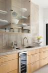 Wet bar featuring light-toned wood cabinetry, stone slab backsplash and countertop, under-counter wine refrigerator, and upper glass-front cabinetry w