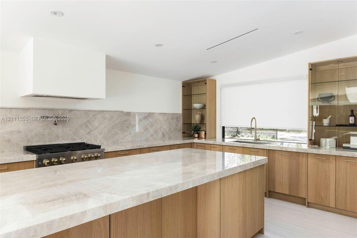 Kitchen featuring natural wood cabinetry, stone-surface countertops and backsplash, integrated glass-front display shelving, wall-mounted pot filler, 