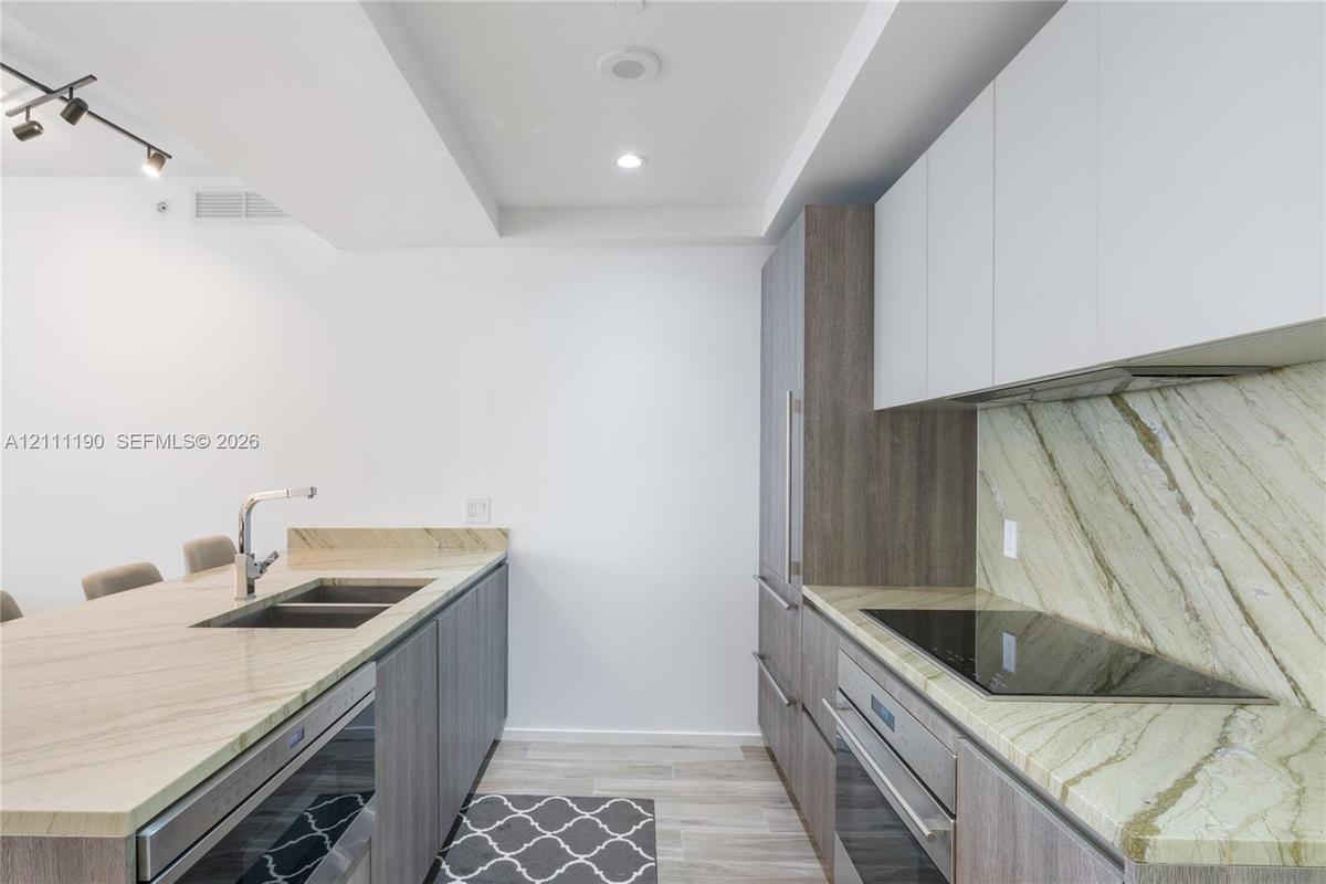 Kitchen featuring stone countertops, stone backsplash, flat-panel wood-finish cabinetry, stainless steel sink with pull-down faucet, and built-in indu