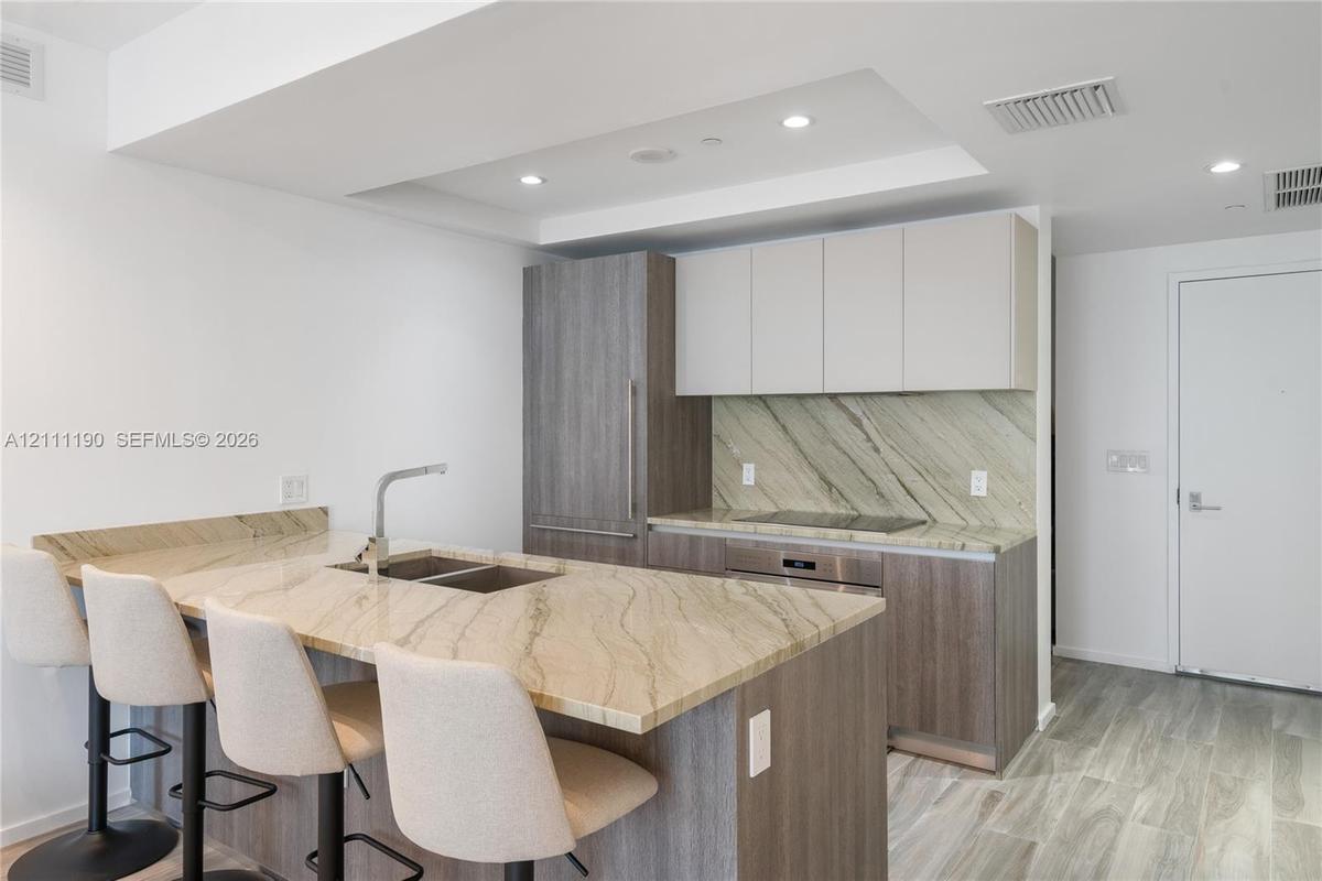 Kitchen featuring two-tone flat-panel cabinetry, stone countertops with matching backsplash, wood-finish flooring, and recessed ceiling lighting