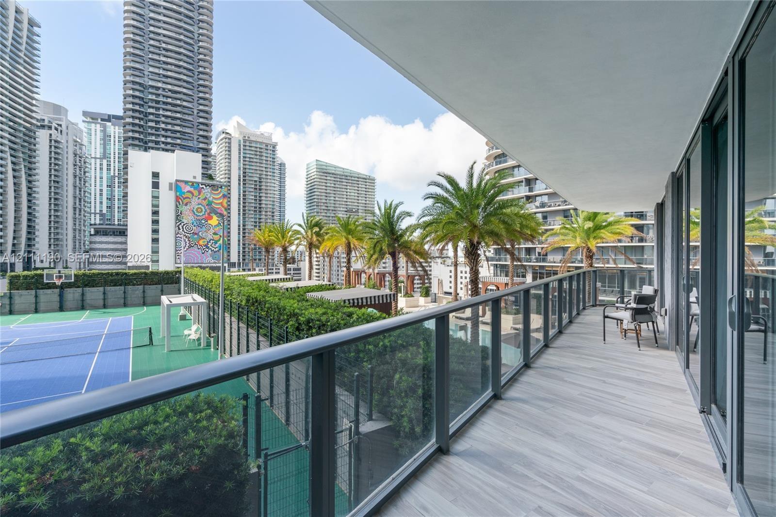 Covered balcony featuring wood-finish tile flooring and a glass panel railing, overlooking a tennis court and urban landscape