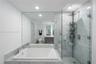 Deep soaking tub with stone tile surround, frameless glass-enclosed shower, expansive wall-mounted mirror, recessed lighting, and stone flooring