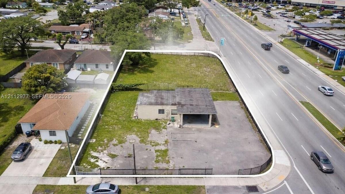 Corner lot featuring a single-story structure with an attached carport, expansive paved surface area, and perimeter chain-link fencing