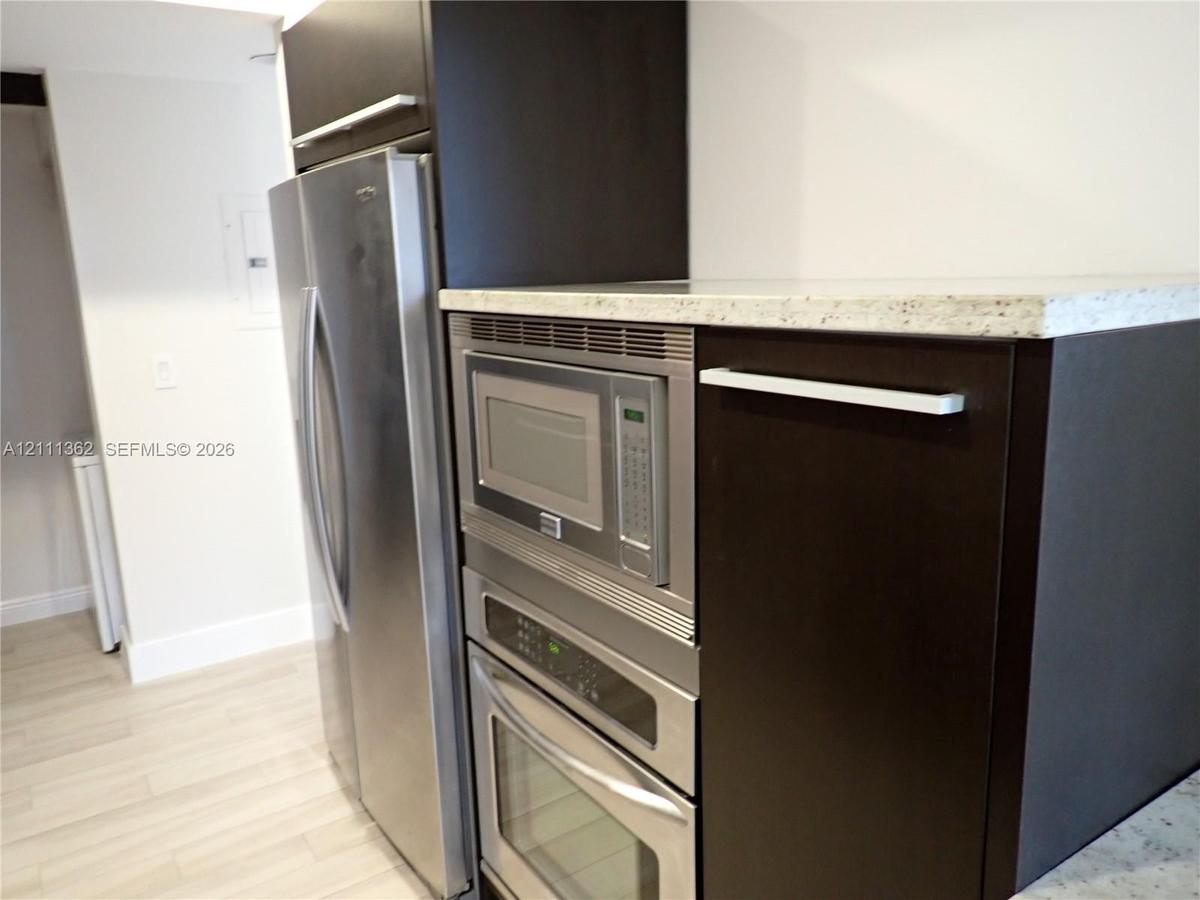 Kitchen featuring dark-finish cabinetry, stainless steel refrigerator, built-in stainless steel microwave and oven, and light-toned stone-finish count