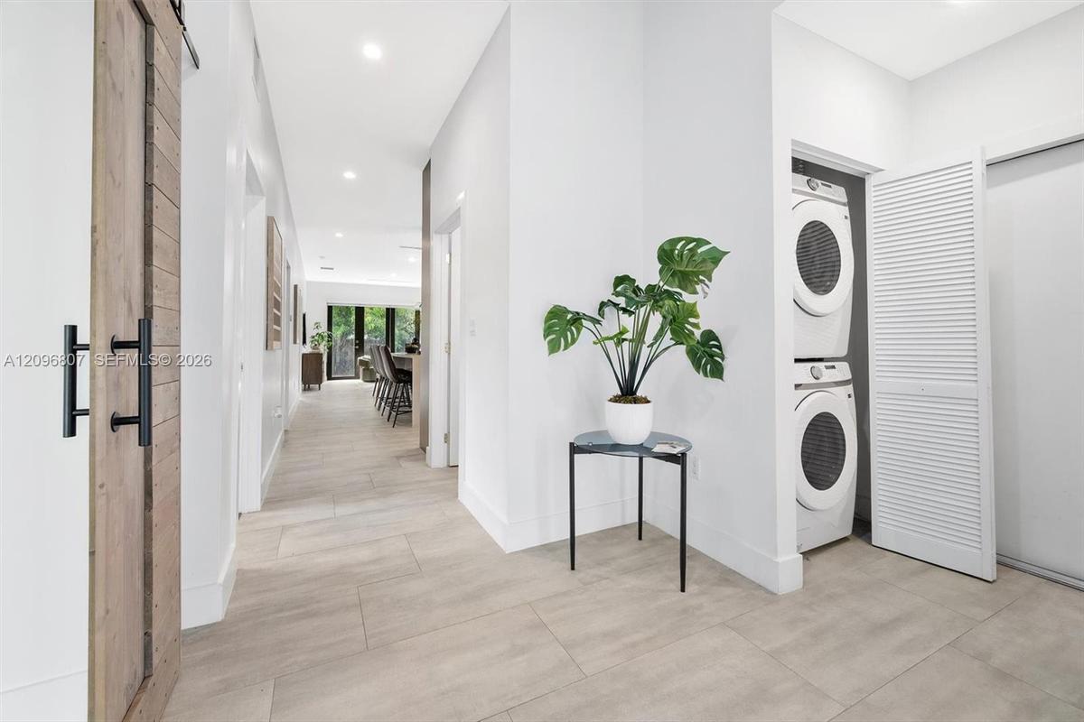 Neutral-tone tile flooring, sliding wood-finish barn door with matte black hardware, and laundry alcove featuring stacked appliances and louvered fold