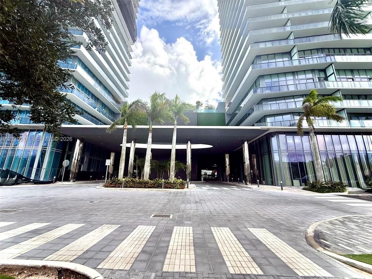 Modern high-rise exterior featuring glass-paneled balconies, floor-to-ceiling windows, a covered porte-cochère, and stone-paved motor court