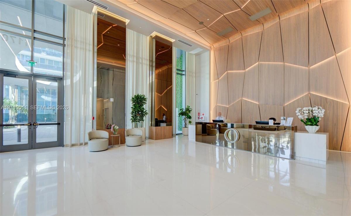 Grand lobby featuring polished white tile flooring, floor-to-ceiling glass entryway doors, geometric wood-paneled walls with integrated recessed light