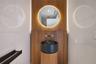 Wall-mounted wood-finish paneling, circular backlit mirror, cylindrical wood-finish pedestal vanity, black stone vessel sink, and wall-mounted metalli
