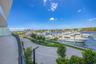 Curved balcony with glass panel railing, paved waterfront walkway, and mature landscaping overlooking a boat marina