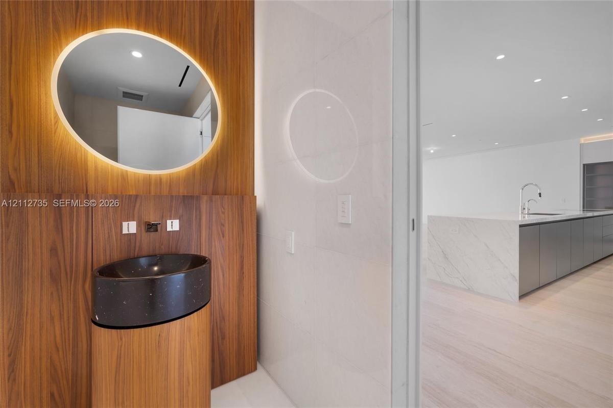 Wood-paneled wall feature with integrated backlit oval mirror, dark speckled pedestal basin, wall-mounted faucet, and stone-surfaced kitchen island wi
