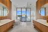 Dual floating wood-finish vanities with vessel sinks and integrated backlit mirrors, freestanding soaking tub, expansive stone-tiled floor, and a glas
