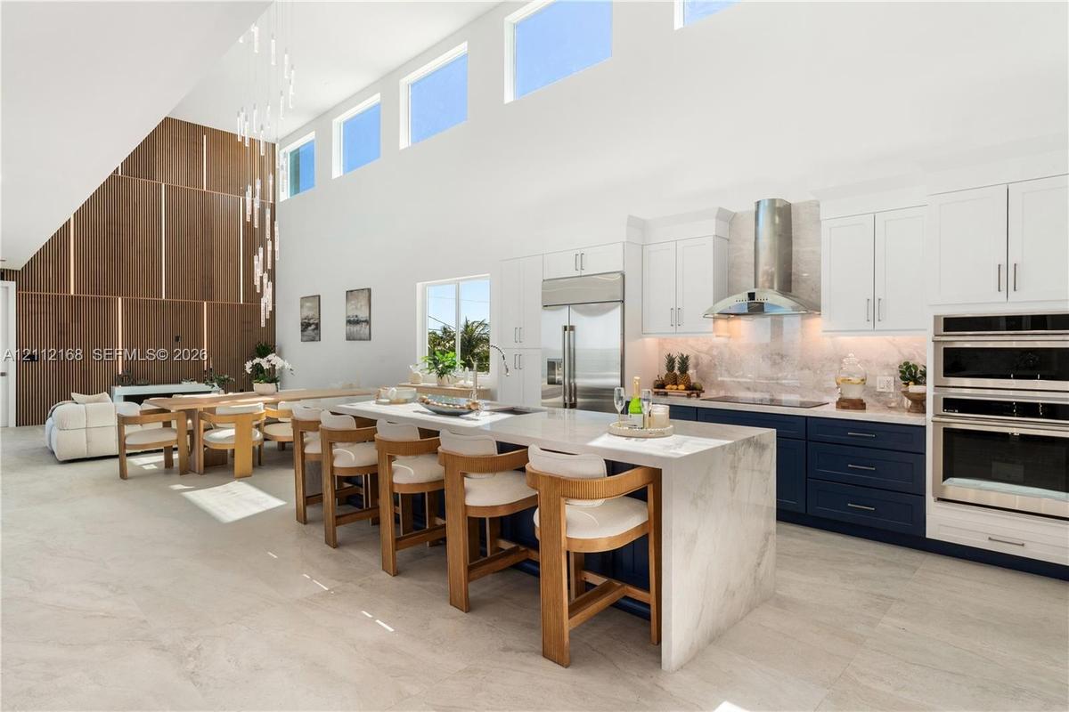 Open-concept kitchen featuring double-height ceilings, a large stone-topped waterfall island, custom two-toned cabinetry, built-in stainless steel app