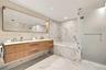 Floating wood-finish vanity with dual sinks, large integrated mirror, walk-in shower with frosted glass enclosure, and stone-tile flooring and wall su
