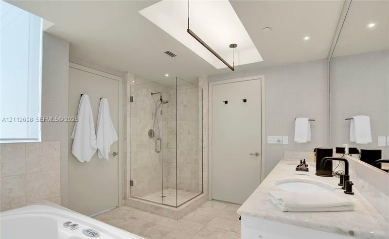 Bathroom featuring a skylight, walk-in glass-enclosed shower with stone tile surround, dual-sink vanity with light stone countertop, and deep soaking 