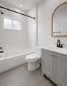 Bathroom featuring a grey vanity with a white countertop, matte black fixtures, an arched wood-frame mirror, tile shower surround, and grey flooring