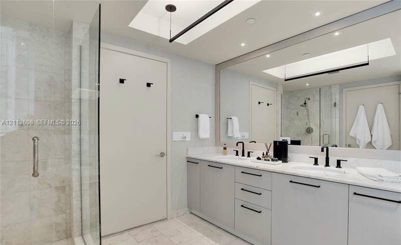 Dual-sink vanity with light-gray cabinetry, stone countertops, matte black fixtures, full-width mirror, and walk-in tiled shower with a glass enclosur