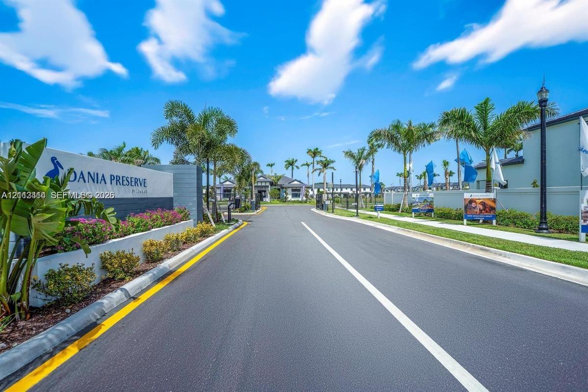 Gated community entrance featuring landscaped beds, mature palm trees, paved roadway, and decorative monument signage