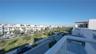 Elevated balcony featuring a white metal railing and a concrete deck surface, overlooking a paved street with green spaces and palm trees