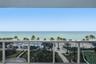 Covered balcony featuring white metal railings, overlooking an oceanfront landscape with a swimming pool, paved walkways, and palm trees