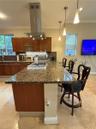 Kitchen featuring a sprawling stone-topped island with a built-in cooktop, stainless steel chimney-style range hood, warm-toned wood cabinetry, and pe