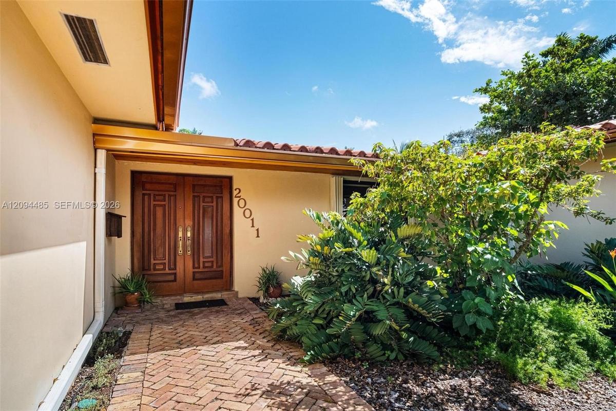 Stucco exterior entry featuring a herringbone-patterned brick walkway, double wood-paneled doors with metallic hardware, and a terracotta tile roof ed