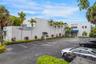 Commercial exterior featuring a concrete facade, expansive asphalt parking area, blue metal awnings, and landscaped perimeter with mature palm trees