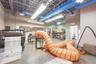 Industrial-style workspace featuring high-clearance exposed ceilings, concrete flooring, linear fluorescent lighting, and extensive blue ductwork