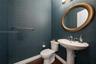 Textured deep-teal wall covering, white pedestal sink with chrome faucet, oval wall-mounted mirror with bronze finish, and dark wood-finish flooring