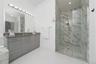 Bathroom featuring a dual-sink vanity with grey cabinetry, a solid-surface countertop, full-width mirror with multi-bulb lighting, and a glass-enclose