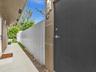 Exterior walkway featuring concrete paving, a white metal privacy fence, stucco siding, and a dark-toned solid entry door with integrated wall-mounted