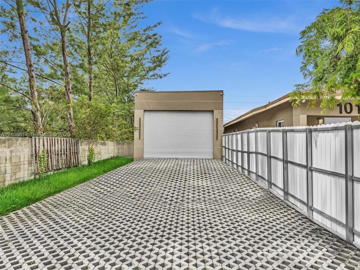 Detached garage with a roll-up door, featuring a permeable paver driveway, white vinyl fencing, and a concrete block perimeter wall