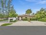 Single-story exterior featuring a stucco facade, covered entryway, concrete driveway, white vinyl fencing, and mature palm landscaping