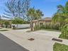 Exterior facade featuring a neutral-toned finish, shingled roof, concrete driveway, white privacy fencing, and mature palm trees