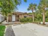 Single-story exterior featuring a covered entryway, concrete paved parking area, perimeter landscaping, and mature palm trees