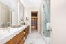 Dual-sink vanity with wood-finish cabinetry, stone-surfaced countertop, full-height backlit mirrors, and glass-enclosed shower with large-format tile 