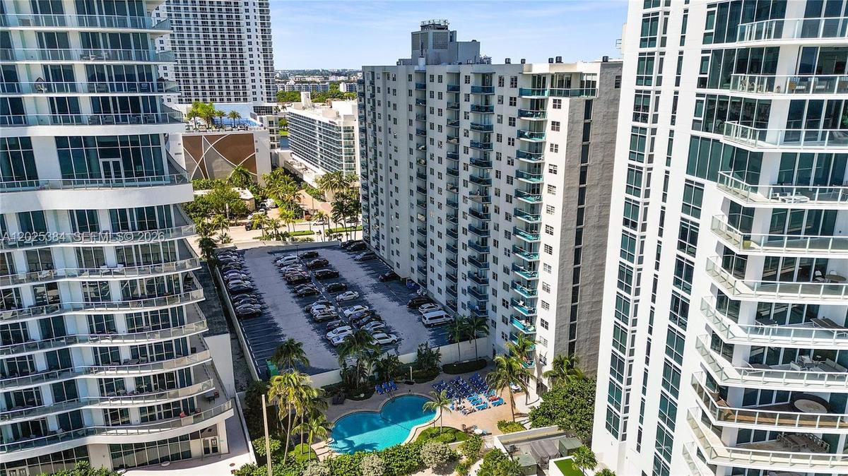 High-rise residential exterior featuring expansive glass balconies, surface parking, and a ground-level swimming pool surrounded by mature palm trees