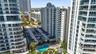 High-rise residential exterior featuring expansive glass balconies, surface parking, and a ground-level swimming pool surrounded by mature palm trees