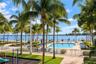 Community swimming pool with a paved deck, manicured green space, and mature palm trees, featuring a gated perimeter and water frontage