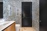 High-contrast patterned wall tile, vanity with a rectangular sink, light-toned countertop, black louvered door, and large-format neutral tile flooring