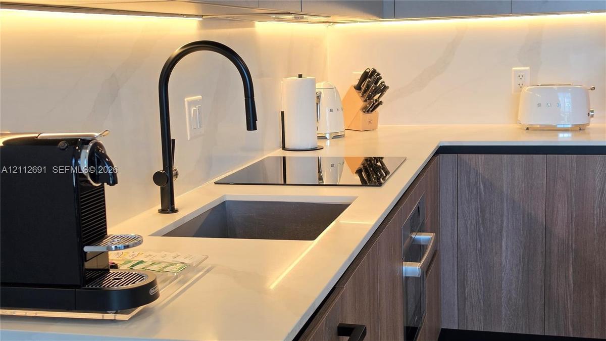 Kitchen featuring light-toned countertops, integrated under-cabinet lighting, wood-finish cabinetry, a square undermount sink, and a matte black high-