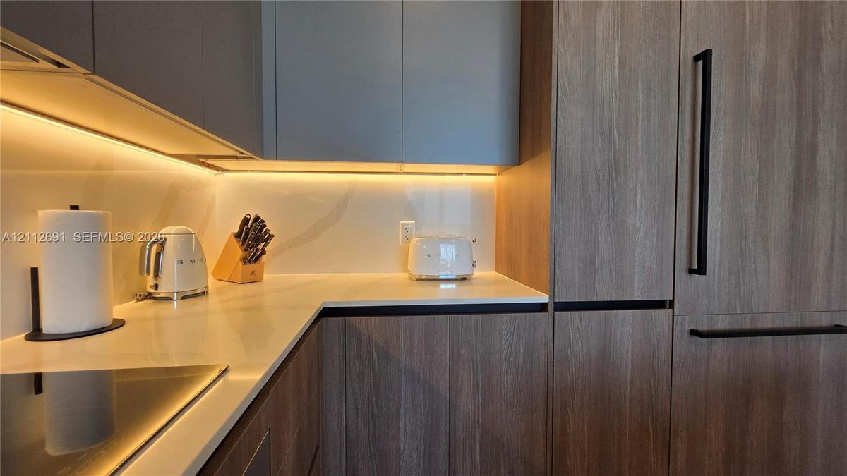 Kitchen featuring wood-finish cabinetry, solid surface countertops, a full-height backsplash, under-cabinet lighting, and integrated panel-ready appli