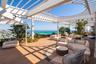 Expansive rooftop terrace featuring a white pergola structure with a circular cutout, wood-finish decking, glass panel railings, and perimeter landsca