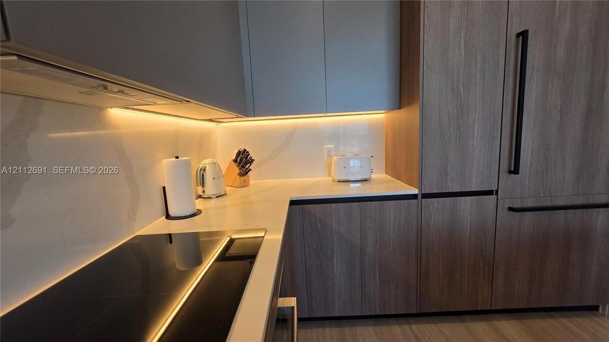 Modern kitchen featuring flat-panel cabinetry with wood-finish grain, integrated paneled refrigerator, solid-surface backsplash, under-cabinet lightin