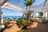 Expansive terrace featuring a circular architectural pergola, wood-finish decking, integrated landscape planters with palm trees, and a glass panel pe