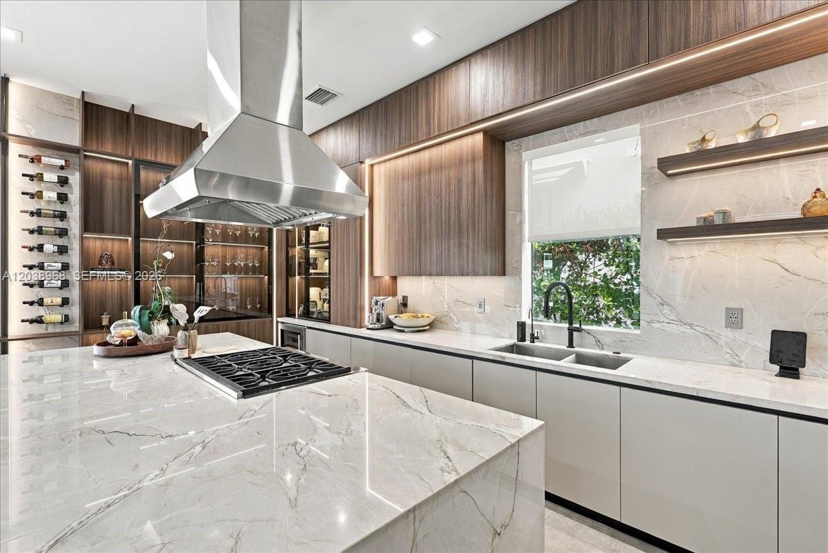 Kitchen featuring a large island with a surface-mounted cooktop, stainless steel vent hood, floor-to-ceiling wood-finish cabinetry, stone-patterned ba