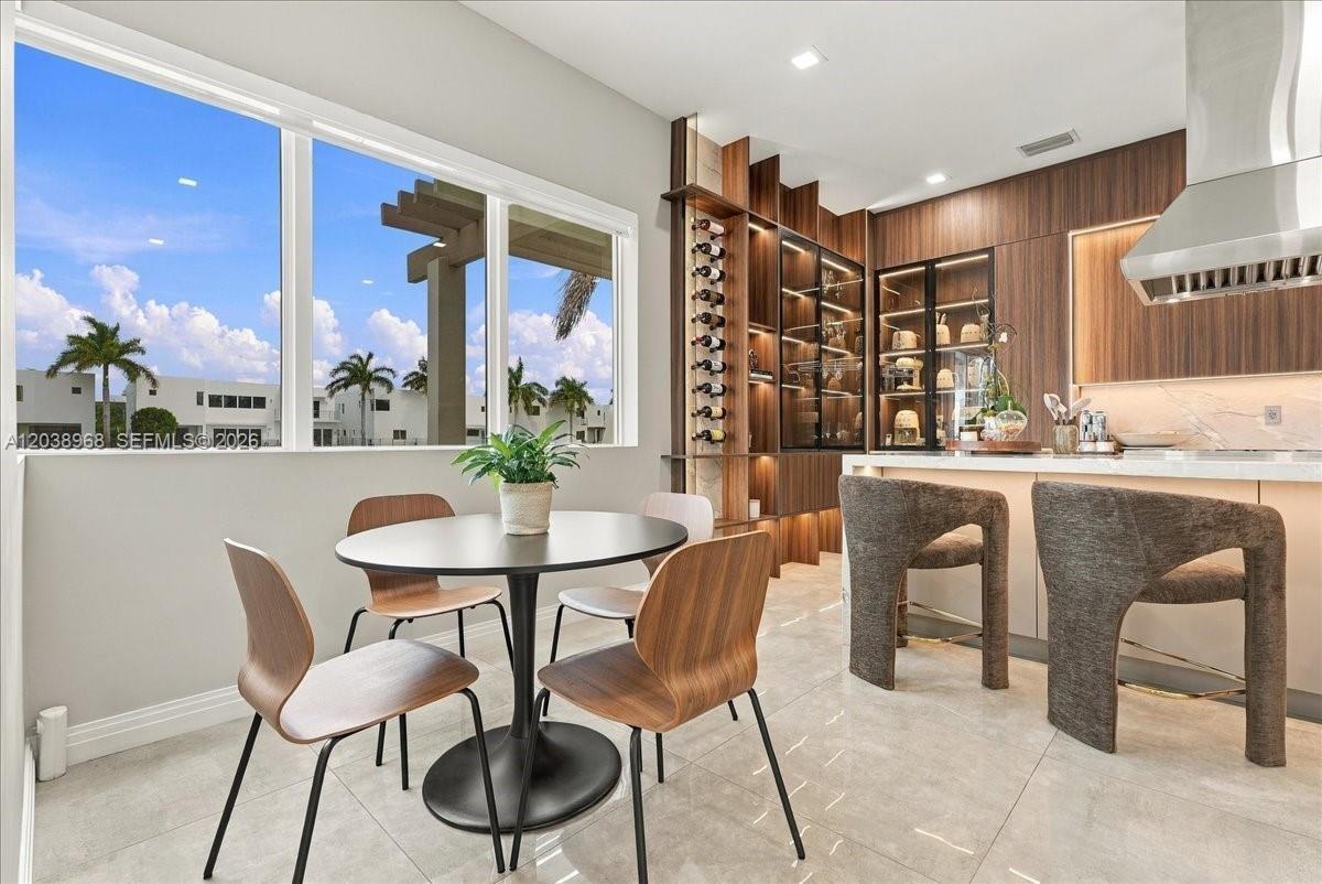 Large windows featuring exterior palm tree and pergola scenery, custom wood-finish cabinetry with integrated wine storage, glass-front display shelvin