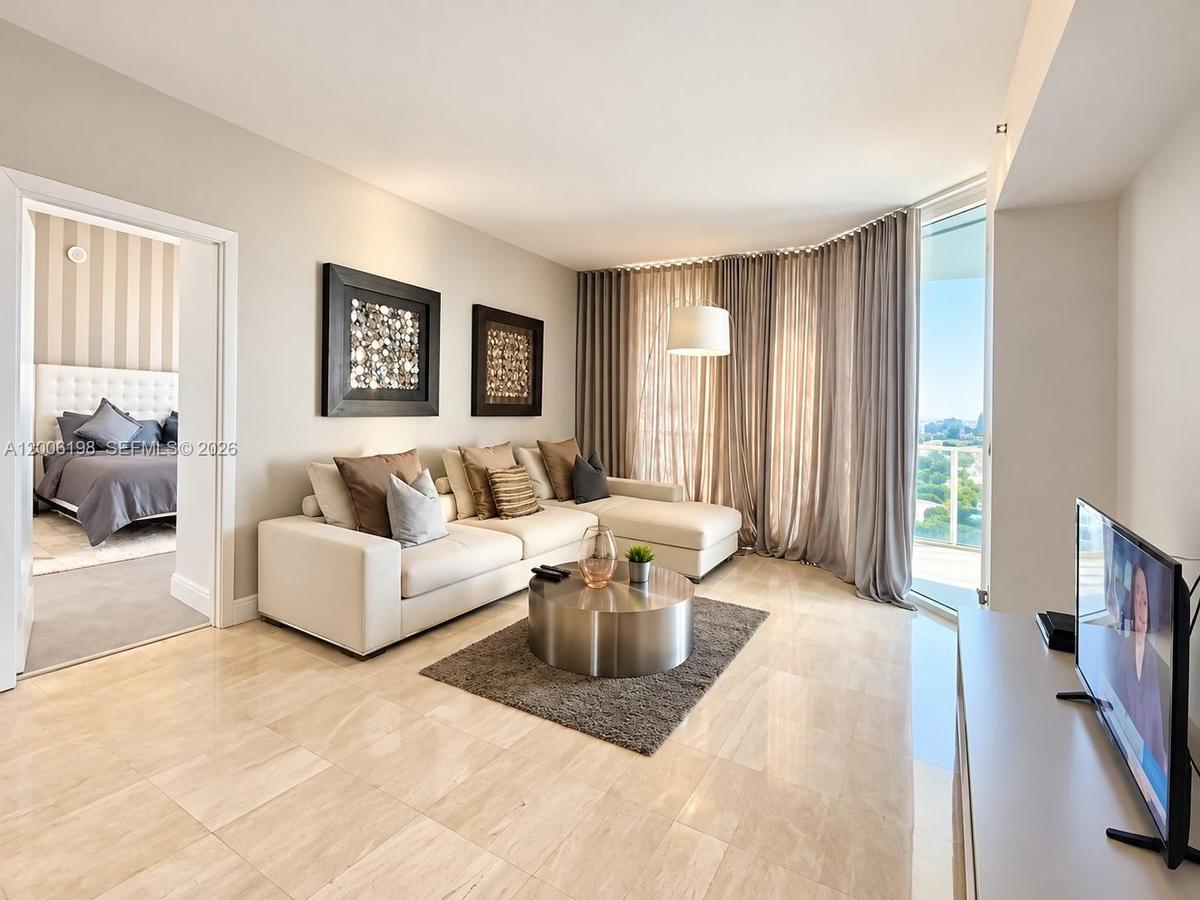 Living area featuring light-toned stone tile flooring, floor-to-ceiling windows, and a glass-paneled balcony