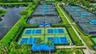 Multiple outdoor pickleball courts with blue surfacing, fenced tennis courts, perimeter landscaping, and waterfront grounds