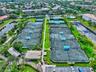 Aerial perspective of a tennis complex featuring multiple hard-surface courts, fenced perimeters, integrated court lighting, and a central clubhouse s